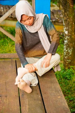 Asyalı Müslüman kadın tahta zeminde bağdaş kurarak oturur, doğal bir şekilde ayaklarına masaj yaparken bir sütuna yaslanır..