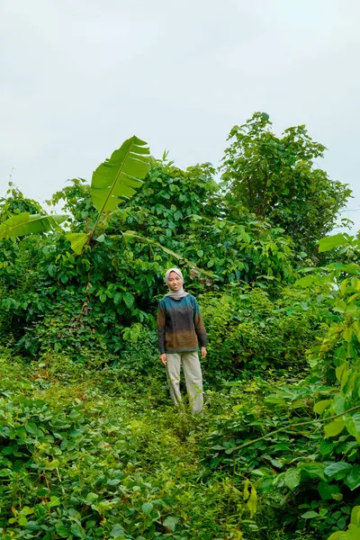 Asyalı Müslüman kadın yemyeşil bitkilerin ortasında dikiliyor ve çevresini gözlemliyor..