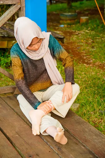 Asyalı Müslüman kadın tahta zeminde bağdaş kurarak oturur, doğal bir şekilde ayaklarına masaj yaparken bir sütuna yaslanır..