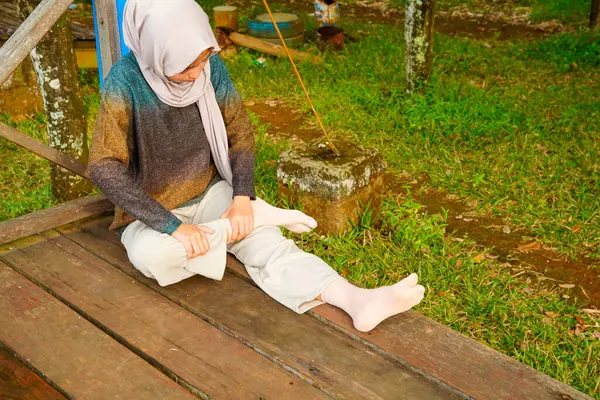 Asyalı Müslüman kadın tahta zeminde bağdaş kurarak oturur, doğal bir şekilde ayaklarına masaj yaparken bir sütuna yaslanır..