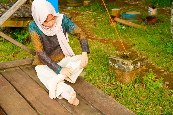 Asyalı Müslüman kadın tahta zeminde bağdaş kurarak oturur, doğal bir şekilde ayaklarına masaj yaparken bir sütuna yaslanır..