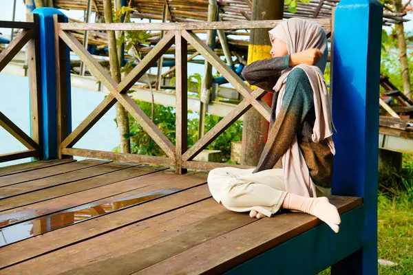 Asyalı Müslüman kadın tahta zeminde bağdaş kurarak oturur, doğal bir şekilde omuzlarına masaj yaparken bir sütuna yaslanır..