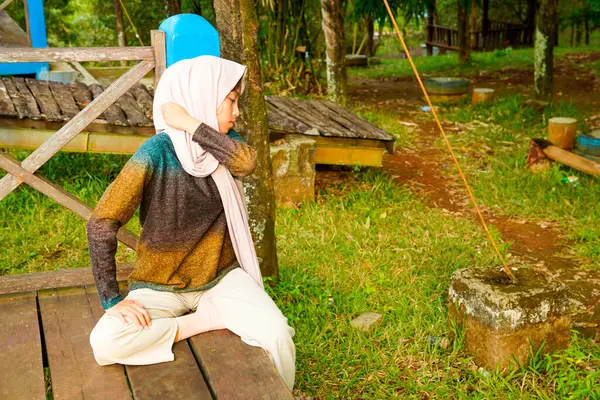 Asyalı Müslüman kadın tahta zeminde bağdaş kurarak oturur, doğal bir şekilde omuzlarına masaj yaparken bir sütuna yaslanır..