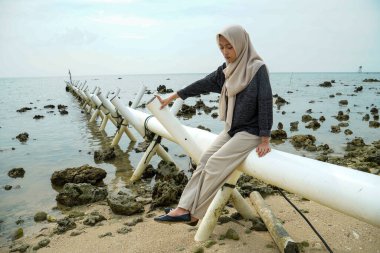 Asyalı Müslüman bir kadının denize açılan bir drenaj borusunda oturduğu görüntüsü kayalık kıyı şeridi ve sığ deniz suyuyla..