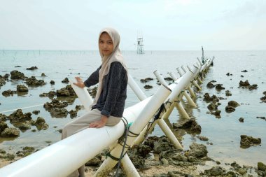 Asyalı Müslüman bir kadının denize açılan bir drenaj borusunda oturduğu görüntüsü kayalık kıyı şeridi ve sığ deniz suyuyla..