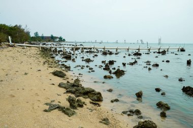 A view of the beach at low tide, with sand dotted with coral reefs and a long waste disposal structure protruding from the shallow waters under a cloudy sky.