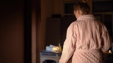 Dövmeli ve bornozlu güzel bir genç kız masaj prosedürüne başlamadan önce vücuduna ve tenine dokunuyor. Mumlu koyu renk güzellik salonu, romantik atmosfer.