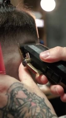 Kolunda dövme olan bir berber berber dükkanında esmer bir adamın saçını kesiyor, kırpma makinesiyle saçını kesiyor. Yüksek kaliteli görüntüler