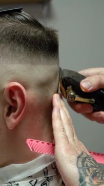 Kolunda dövme olan bir berber berber dükkanında esmer bir adamın saçını kesiyor, kırpma makinesiyle saçını kesiyor. Yüksek kaliteli görüntüler