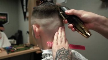Kolunda dövme olan bir berber berber dükkanında esmer bir adamın saçını kesiyor, kırpma makinesiyle saçını kesiyor. Yüksek kaliteli görüntüler