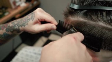 Kolunda dövme olan bir berber berber dükkanında esmer bir adamın saçını kesiyor, kırpma makinesiyle saçını kesiyor. Yüksek kaliteli görüntüler