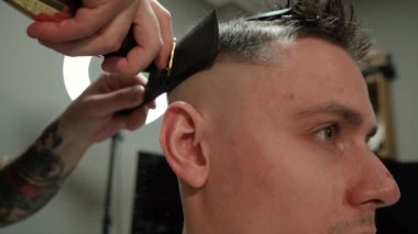 Kolunda dövme olan bir berber berber dükkanında esmer bir adamın saçını kesiyor, kırpma makinesiyle saçını kesiyor. Yüksek kaliteli görüntüler