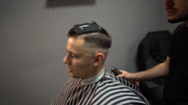 Kolunda dövme olan bir berber berber dükkanında esmer bir adamın saçını kesiyor, kırpma makinesiyle saçını kesiyor. Yüksek kaliteli görüntüler