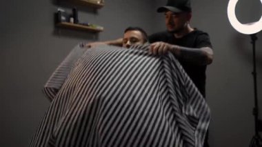 Kolunda dövme olan bir berber berber dükkanında esmer bir adamın saçını kesiyor, kırpma makinesiyle saçını kesiyor. Yüksek kaliteli görüntüler