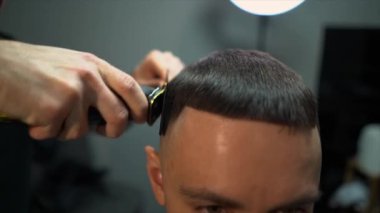 Kolunda dövme olan bir berber berber dükkanında esmer bir adamın saçını kesiyor, kırpma makinesiyle saçını kesiyor. Yüksek kaliteli görüntüler