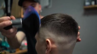 Kolunda dövme olan bir berber berber dükkanında esmer bir adamın saçını kesiyor, kırpma makinesiyle saçını kesiyor. Yüksek kaliteli görüntüler