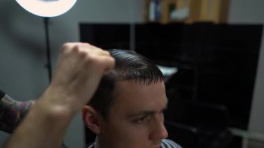 Kolunda dövme olan bir berber berber dükkanında esmer bir adamın saçını kesiyor, kırpma makinesiyle saçını kesiyor. Yüksek kaliteli görüntüler