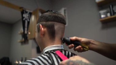 Kolunda dövme olan bir berber berber dükkanında esmer bir adamın saçını kesiyor, kırpma makinesiyle saçını kesiyor. Yüksek kaliteli görüntüler