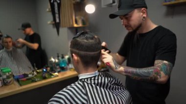 Kolunda dövme olan bir berber berber dükkanında esmer bir adamın saçını kesiyor, kırpma makinesiyle saçını kesiyor. Yüksek kaliteli görüntüler