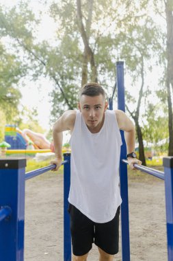 Yakışıklı şişirilmiş genç vücut geliştirici sokakta yatay bar ve paralel barlarda egzersiz yapıyor. Güzel erkek vücut şekilleri, güzellik ve sağlık, eğitim ve motivasyon. Yüksek