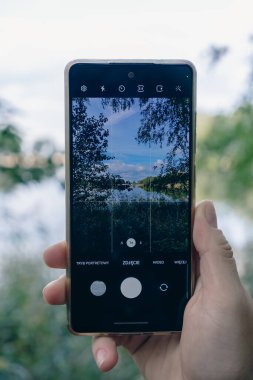 Bir insan ormandaki bir gölün ve doğanın fotoğraflarını cep telefonuyla çeker..