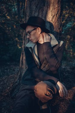Şapkalı ve gözlüklü genç bir adam ağaç gövdesinin yanında oturuyor.
