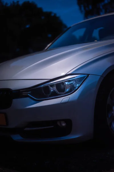 2012 BMW 3 serisi farının detaylı görüntüsü. Gece çekilen bir sokak lambası gümüş arabanın kaputunu ve farını aydınlatıyor..