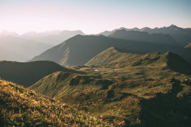 Dağları ve bulutları olan güzel bir manzara. Dağ yamaçlarının panoramik manzarası, çiçek çayırları, yüksek dağ çiçekleri, günbatımında yaz yeşili ve sarı tepeler. Uzakta küçük bir dağ gölü. Yüksek açılı atış. Geniş açı.
