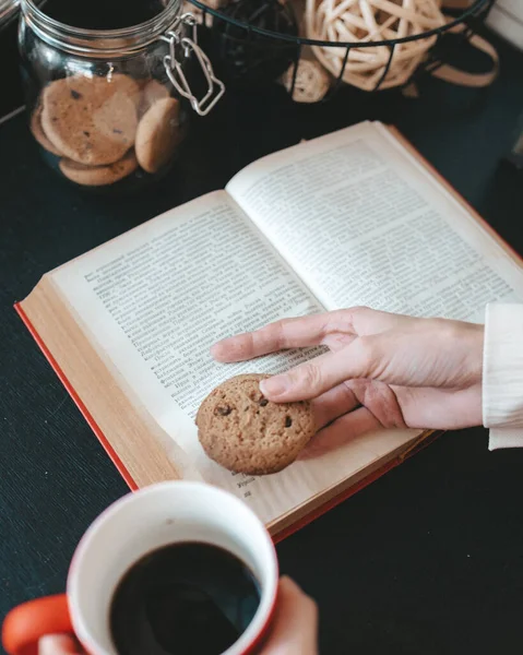 Kahve dükkanında bir fincan kahve ve kurabiyeyle kitap okuyan bir kadın, kitaptaki bir kadın eli, siyah arka plandaki bir kitap, kadın elinde kurabiyeler, kırmızı bardaktaki siyah kahve, çikolatalı kurabiyeler, kahve içen ve okuyan bir kadın, kurabiye kavanozunu kapat.