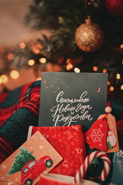 Hediyeler, dekorasyonlar ve bulanık bir arkaplanda süslü bir tebrik kartıyla Noel ağacı. Açık bir hediye, tebrik ederim. Yeni yıl. Kırmızı ve yeşil renkli hediye, yakın plan. Dikey. Rusça bir kartpostal için tebrik mesajı at.