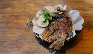 Kahverengi yağ kağıdıyla kaplı tabakta salatalık dilimleri ve fesleğen yapraklarıyla ızgara gourami balığı. Ahşap masa arkaplanlı yatay kopyalama alanının üst görünümü.