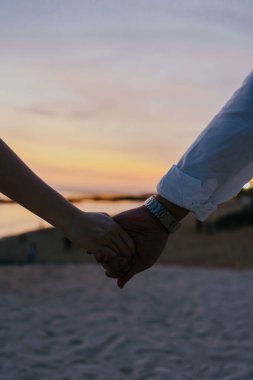 Sahilde el ele tutuşan ve arka planda güzel bir gün batımı olan bir çiftin yakın plan çekimi..