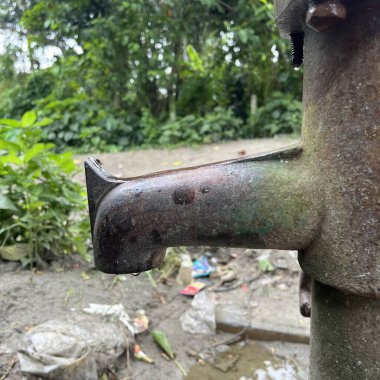 Su damlacıklarıyla dolu yıpranmış bir su pompasının yakın çekimi paslı dokusunu sergiliyor, çamur, çöp ve bulanık kırsal yeşillik zeminine karşı kuruluyor..