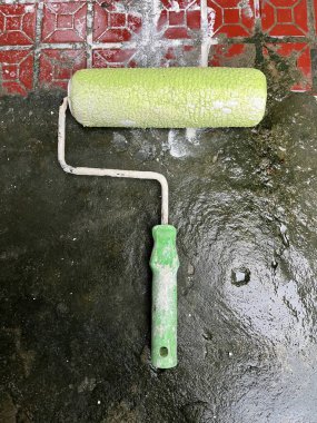 Yukarıdan bakıldığında açık yeşil saplı ve desenli silindirli bir boya silindiri, ıslak, koyu bir zeminde kırmızı fayansların yanında dinleniyor..