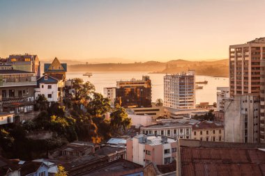 Şili 'nin Valparaiso kentinin panoramik manzarasında, yamaçlara inşa edilmiş geleneksel evler, renkli duvar resimleri, modern kuleler ve demirli gemileri olan bir körfez yer alıyor..