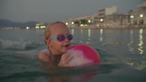 一个6岁的金发女郎 戴着泳镜 高高兴兴地在暮色的大海中游泳 手里拿着一个橡皮球 城市的灯光照亮着它的背景 — 图库视频影像