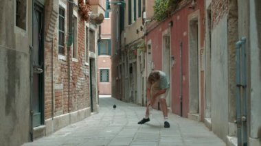 İtalya, Venedik 'te bir sokakta şişe çevirme yarışması düzenleyen bir genç. Şişe dik düşerken ellerini kaldırır ve arka planda bir güvercin uçar.