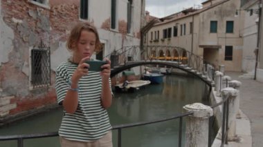 Bir genç Venedik kanalının yakınında akıllı telefon tutuyor. Arka planda bir köprü görülüyor
