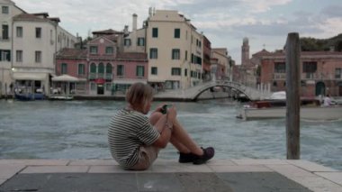 Çizgili tişörtlü bir genç Venedik kanalının kenarında otururken ve arka planda tarihi binalar dururken telefonuyla büyülenir.