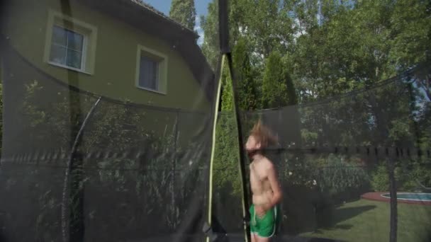 水泳ショートパンツの12歳の少年は 彼の後ろに住宅の庭の設定とトランポリンでジャンプを楽しんでいます — ストック動画