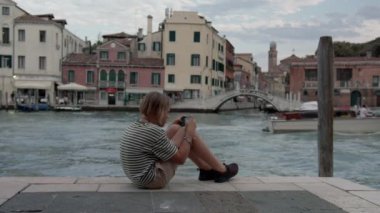 Çizgili tişörtlü bir genç Venedik kanalının kenarında otururken ve arka planda tarihi binalar ve botlarla telefonuyla büyülenir.