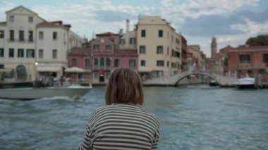 Venedik Kanalı 'ndaki tarihi binaları gözetleyen çizgili tişörtlü bir gencin arka görüntüsü.