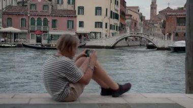 Venedik kanalında kürek çeken bir gondolcuya odaklan, sonra çizgili tişörtlü bir genci yanına çek ve cep telefonuyla oyna.