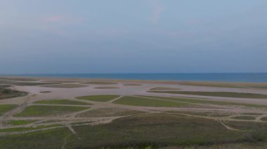 Yükselmiş bir manzara, sakin bir denize bitişik sulak alanların karmaşık desenlerini yakalar. Bitki örtüsü ve kum parçaları, akşamın erken saatlerinde solan ışığın altında doğal bir mozaik yaratır.