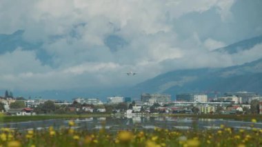 Bir uçak, bir çiçek tarlası boyunca görünen, dağlardan oluşan bir zemin ve bulutlu gökyüzü ile bir şehrin üzerine inmek için yaklaşıyor. Avusturya Alpleri. Innsbruck Havaalanı