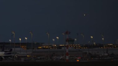 İniş ışıklarıyla dolu bir uçak havaalanı terminaline gece yaklaşırken, arka planda pist ve terminal ışıkları yanıyor.