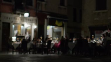 Ziyaretçiler Venedik 'te bir açık hava restoranında yemek yiyorlar, tarihi mimari arasında bulanık ışıklar ve siluetler var.