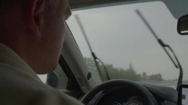Bir adam araba kullanırken tek kullanımlık bardaktan kahve içer, silecekler yağmurlu bir günde açıktır, ve navigasyon sistemi gösterge panelinde görünür.