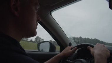 Araba süren bir adamın profili, yağmur yağarken yola dikkatle odaklanmış, silecekleri hareket halinde ve yan camında yağmur damlaları var.