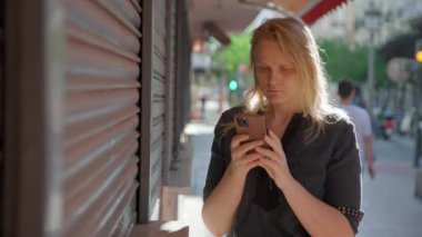 Siyah bluzlu bir kadın kaldırımda dikilirken akıllı telefonuna odaklanmış ve şehir hayatı arka planda bulanıklaşmış.